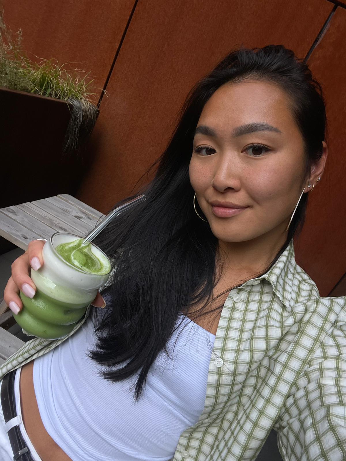 A woman with long black hair is shown in a close-up selfie, holding a green matcha drink. She is wearing a white tank top, a light green and white checkered button-up shirt, and hoop earrings, set against a warm brown wooden wall background.